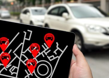 Em primeiro plano, uma mão segurando um ipad e na tela mostrando uma ilustração de geolocalização de carros. No segundo plano, carros.
