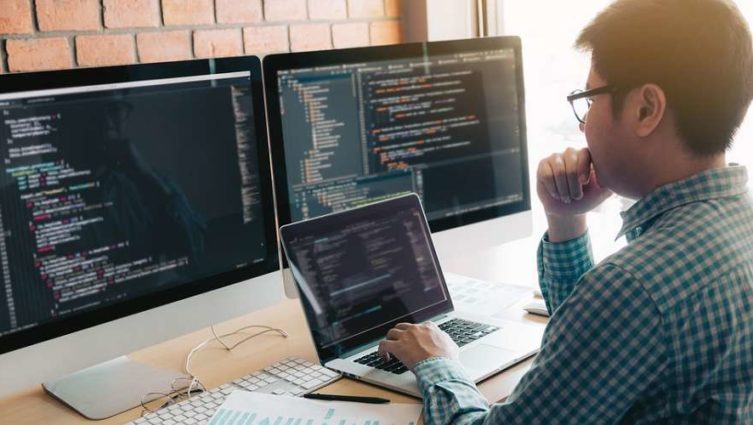 Homem olha para computador e monitores que exibem códigos de programação